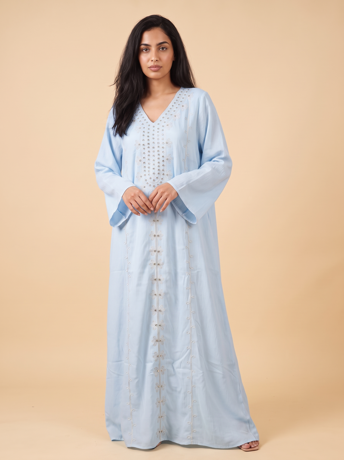 A woman modeling a light blue kaftan dress with silver embroidery and button details, standing against a solid beige studio background.