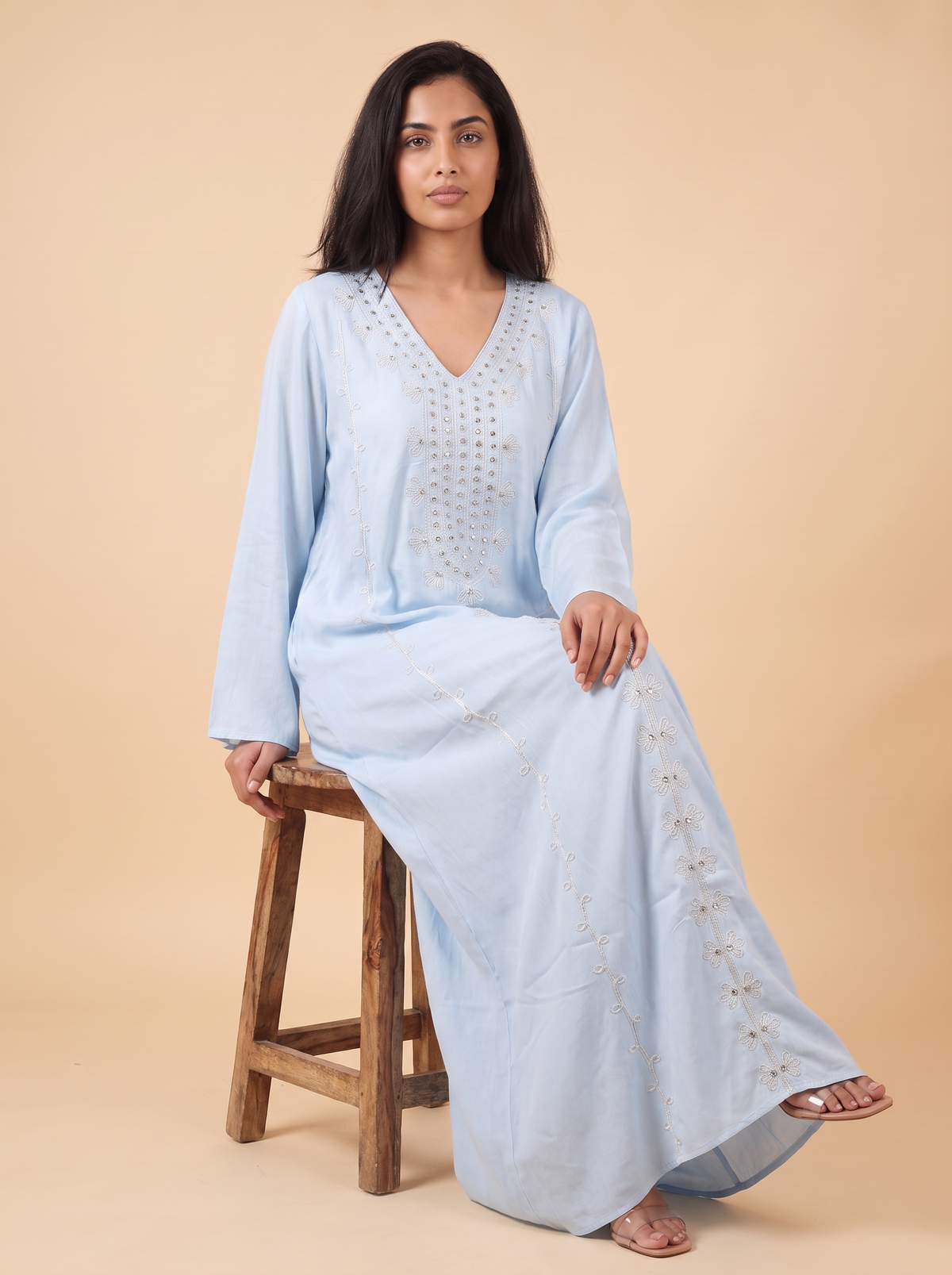 A woman wearing a light blue long kaftan dress with silver embroidery, sitting on a rustic wooden stool against a solid beige background.