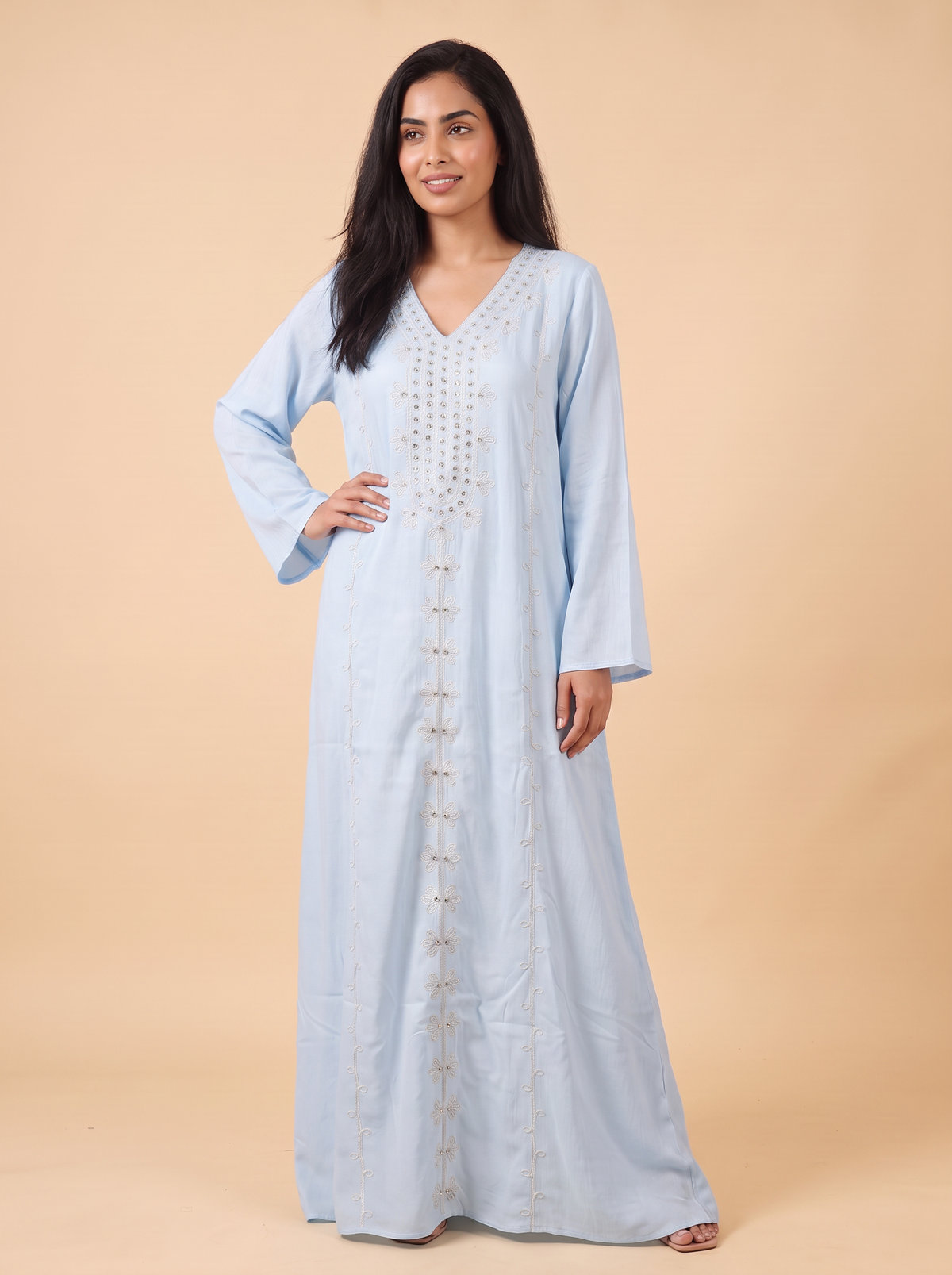 A woman modeling a light blue kaftan dress with gold embroidery and button details, standing against a solid beige background.