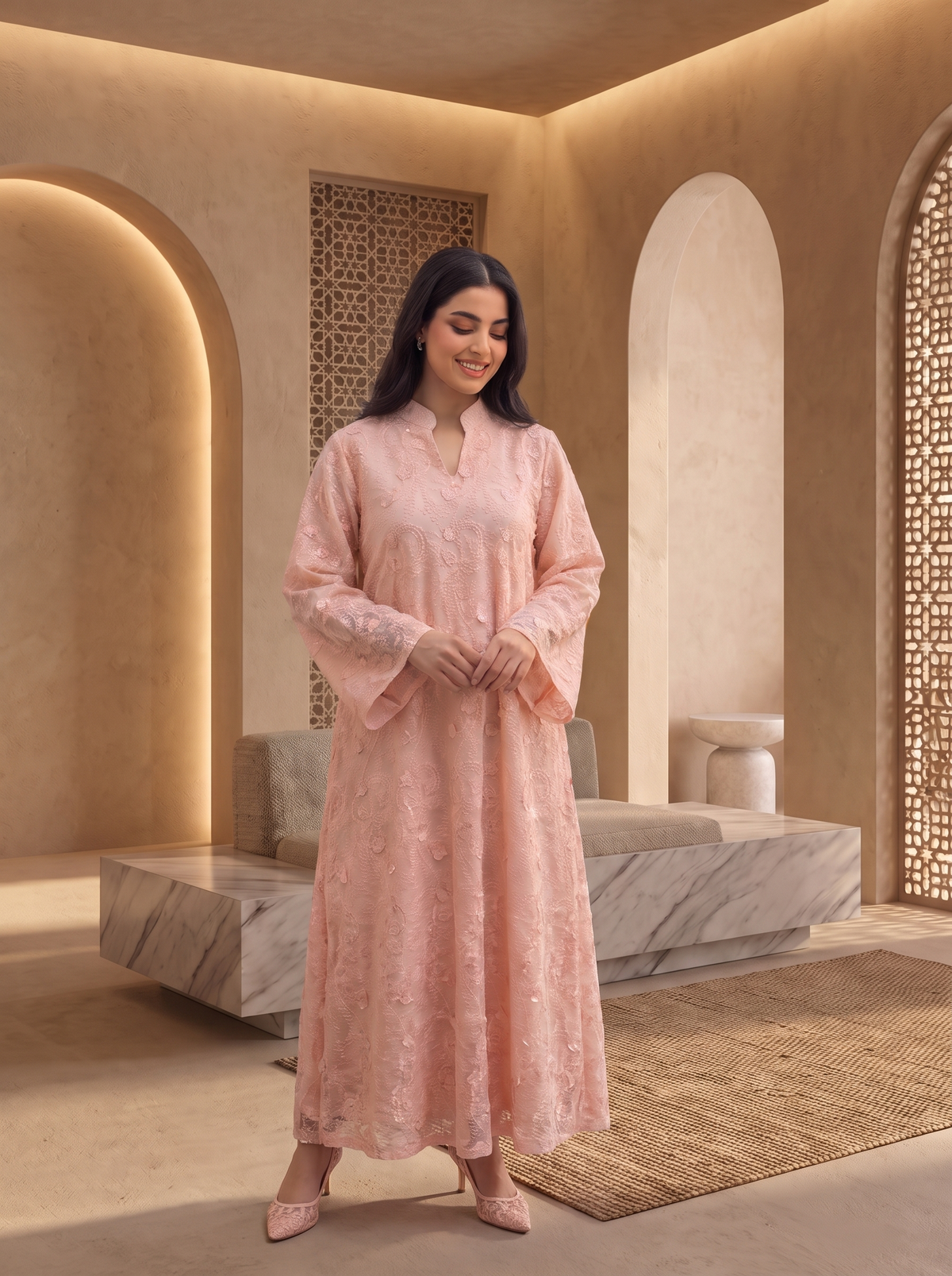A woman models a light pink embroidered kurta set in a warm, modern architectural setting with beige walls and arches. The set includes a long-sleeved tunic with wide flared sleeves and a matching long skirt.