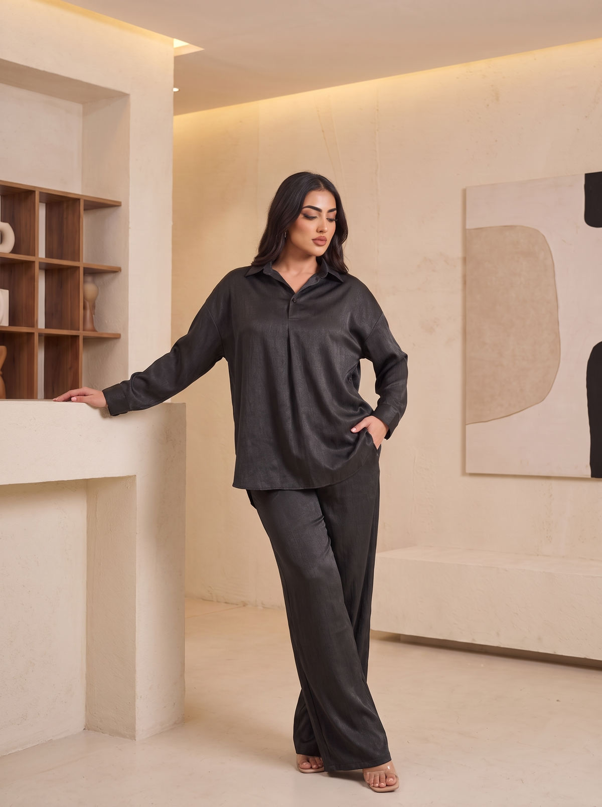 A woman modeling a dark gray two-piece pajama set consisting of a long-sleeve collared button-up shirt and matching wide-leg pants, standing in a modern minimalist interior with warm beige walls and shelves.