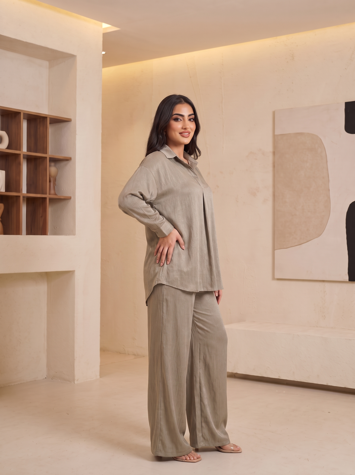A woman modeling a matching two-piece lounge set in a warm, modern beige interior. The set consists of a long-sleeve button-up shirt and wide-leg trousers in a light taupe-beige linen-like fabric.