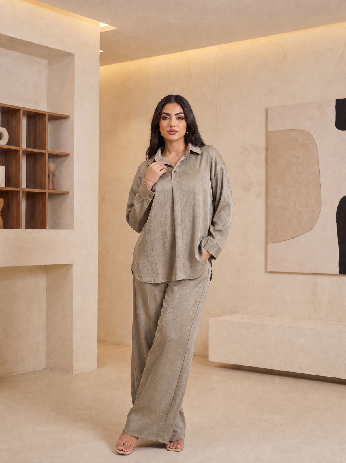 A woman modeling a matching two-piece lounge set in a warm, modern beige interior. The set consists of a long-sleeve button-up shirt and wide-leg trousers in a textured, light taupe fabric.