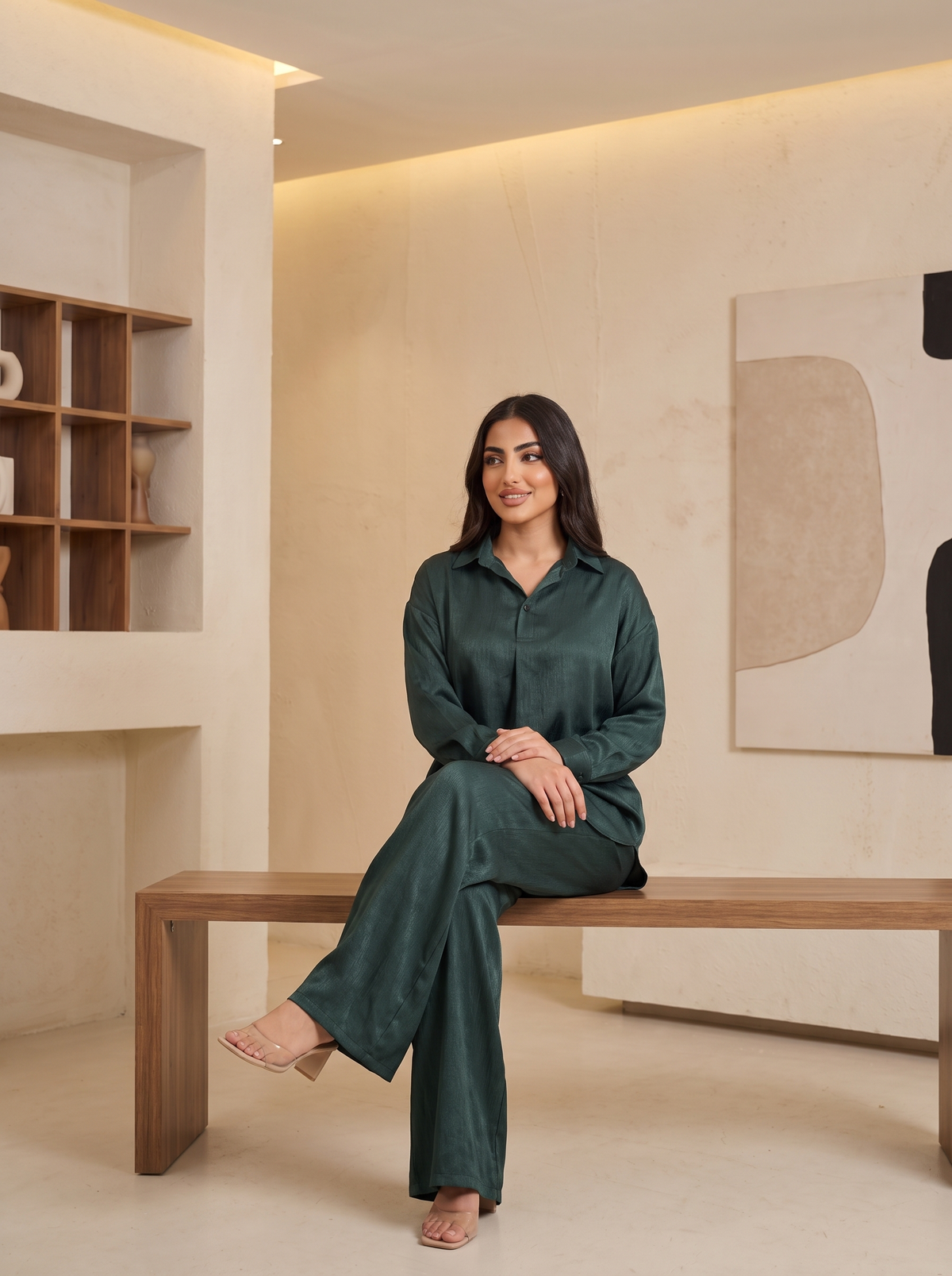 A woman wearing a dark green satin pajama set sits on a wooden bench in a modern, minimalist room with warm beige walls and built-in shelving.