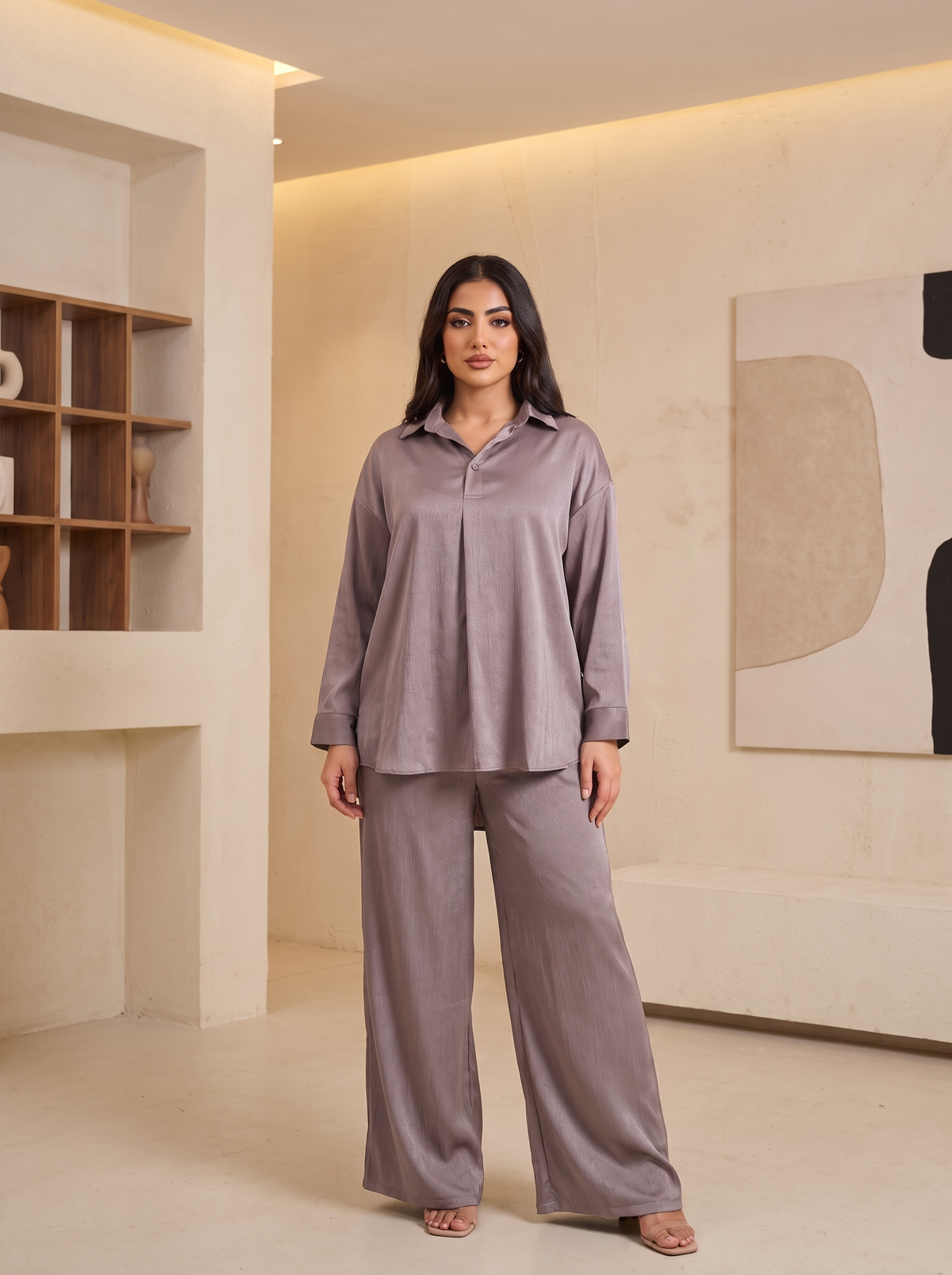 A woman modeling a matching two-piece lounge set in a modern beige-toned interior. The set consists of a long-sleeve button-front top and wide-leg pants in a muted dusty mauve or taupe color.