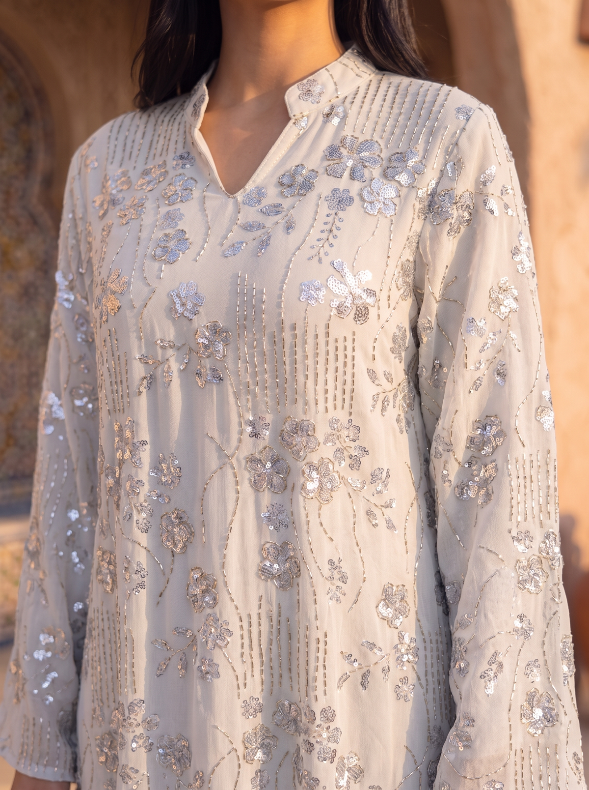 A close-up of a woman wearing a light beige or off-white long kurta with intricate silver and white floral embroidery and sequin embellishments, set against a warm, blurred architectural background.