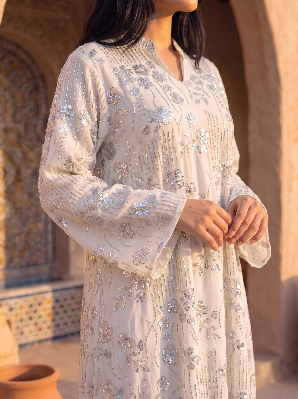A woman wearing a long-sleeved cream-colored embroidered tunic with silver and gold sequins, standing in front of a warm-toned archway.