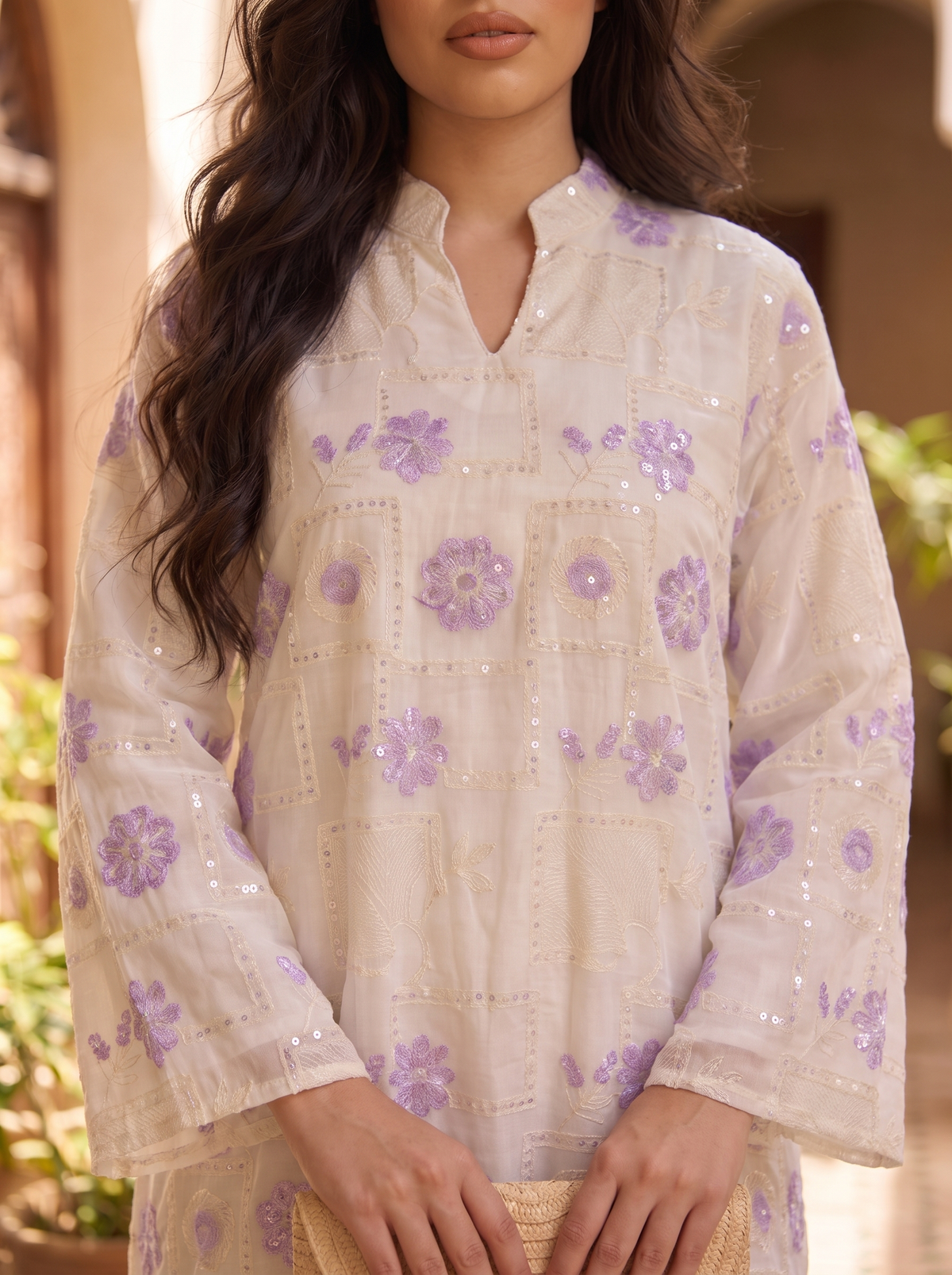 A woman wearing a cream-colored embroidered tunic top with purple floral patterns and sequins, photographed in a warm, softly blurred outdoor setting.