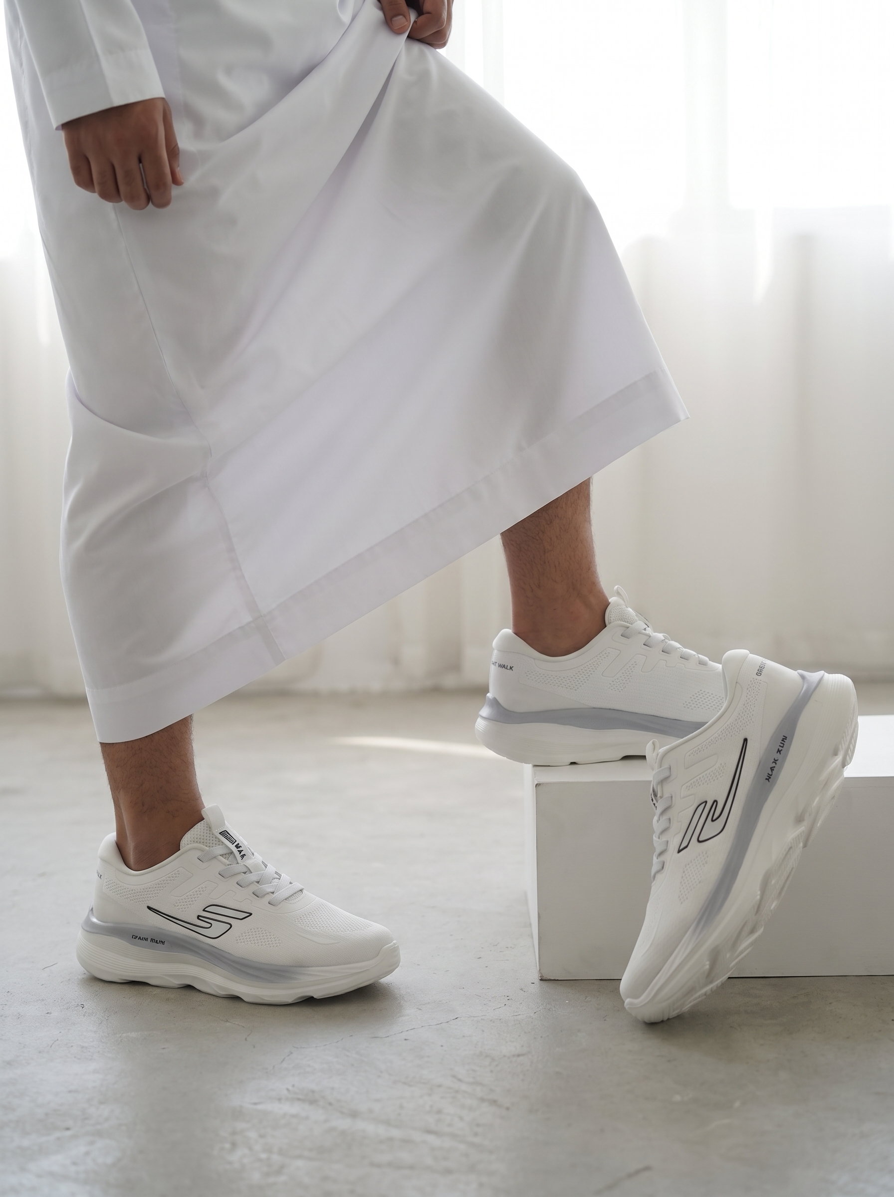 A person wearing white sneakers with a long white robe, standing on a concrete floor with one shoe resting on a white block. The sneakers have a chunky white sole with a light gray accent stripe and a stylized gray logo on the side.