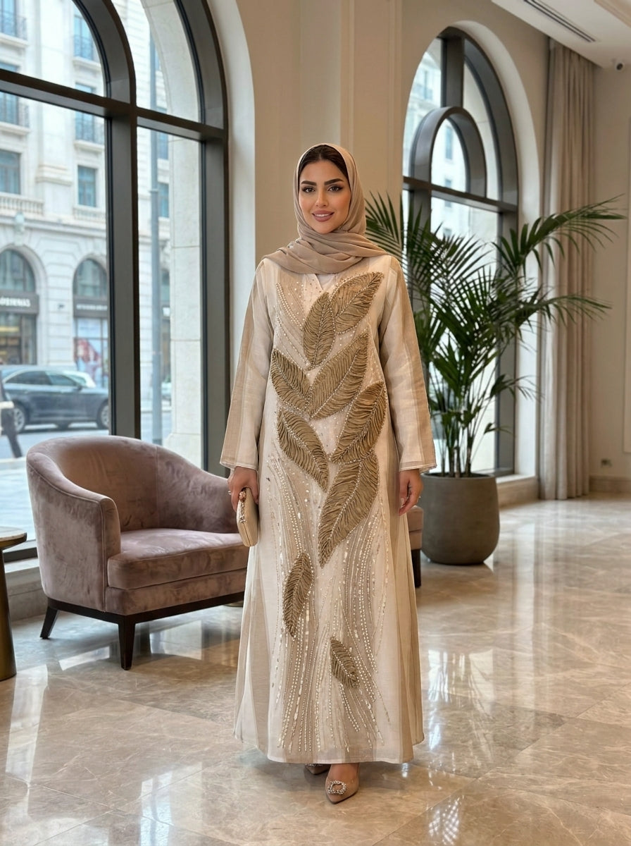 Woman in a beige and gold dress standing in a stylish interior setting with large windows and a plant.